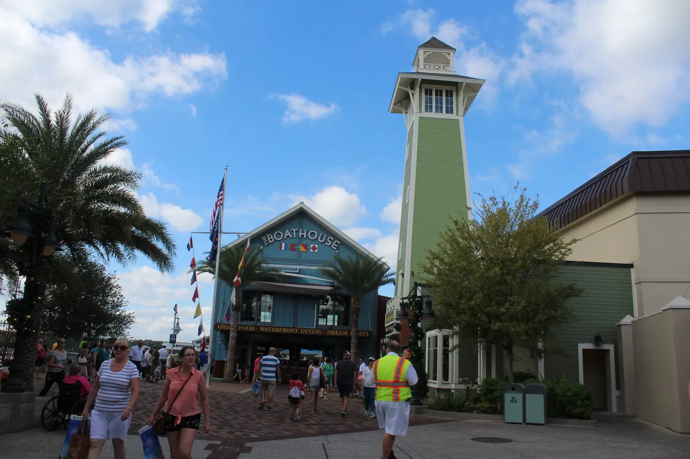 The Boathouse restaurant's blue waterfront building with nautical flags and a tall green lighthouse-style tower at Disney Springs, with visitors walking past