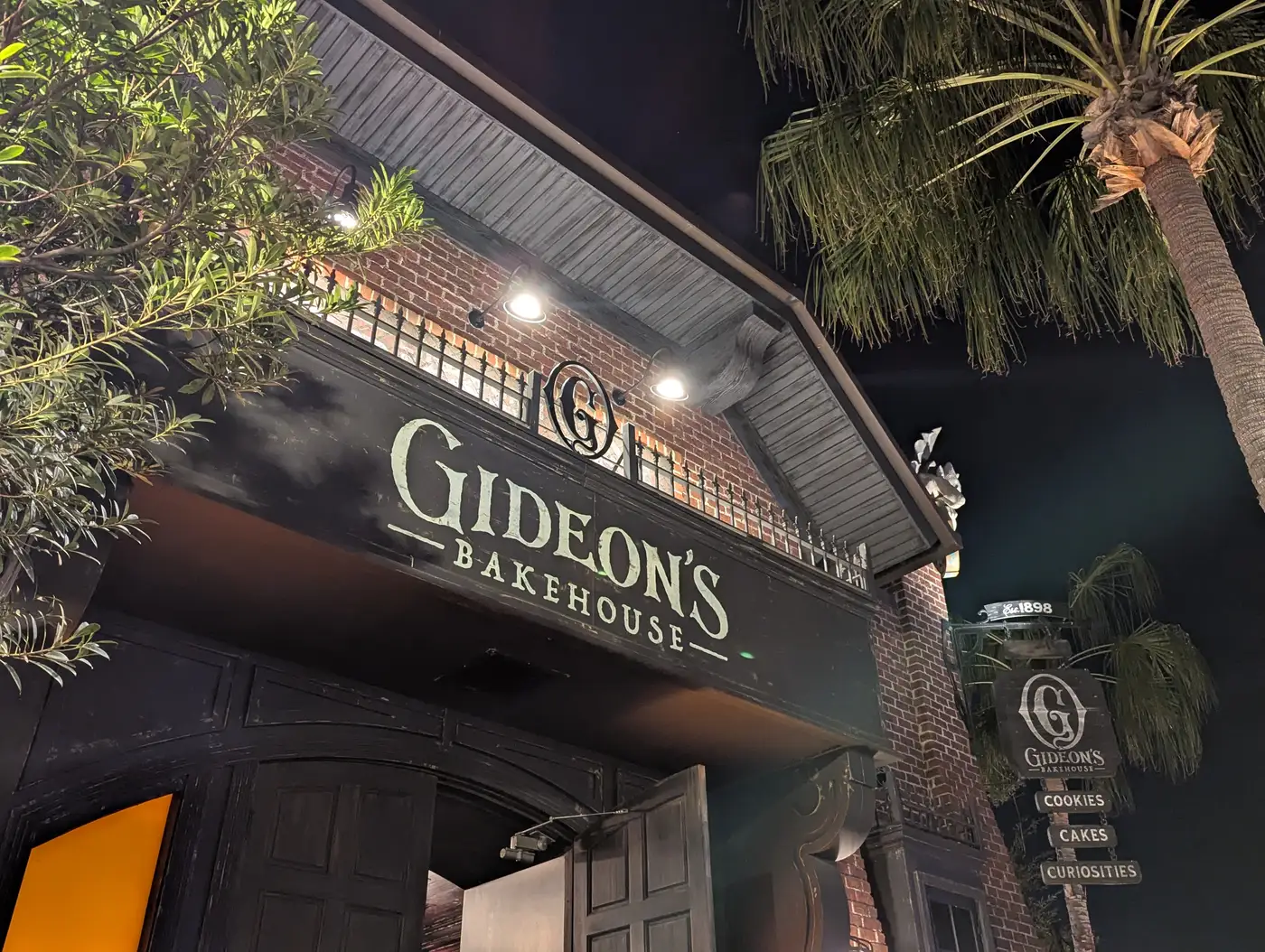 The Gideon's Bakehouse sign over its entrance at Disney Springs at night, brick facade lit from below with a palm tree beside it
