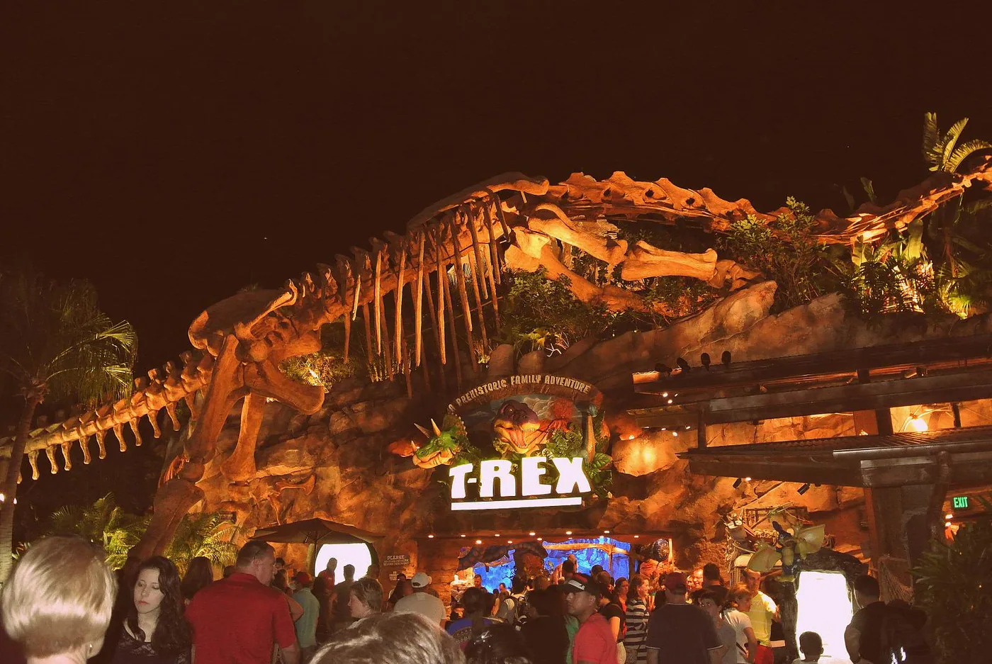 T-REX restaurant's illuminated sign at night beneath a giant dinosaur skeleton sculpture, with a 'Prehistoric Family Adventure' tagline, at Disney Springs