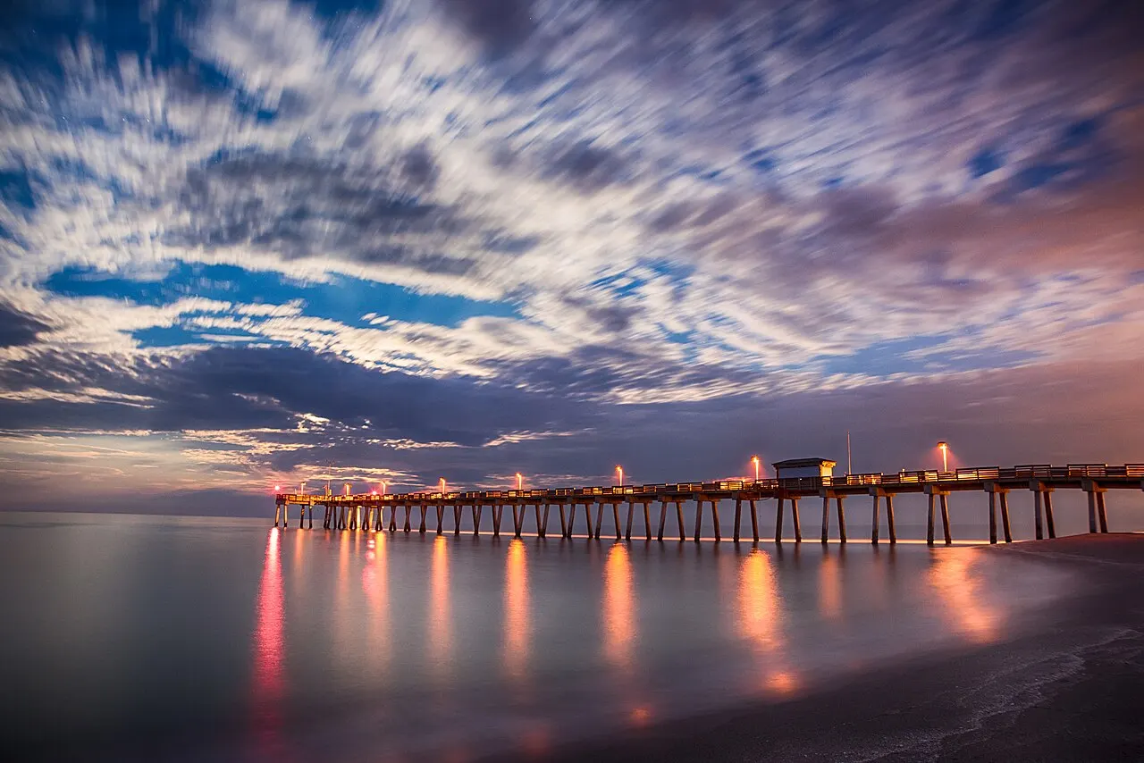 The Venice Fishing Pier at dusk, silhouetted against a dramatic blue and orange sky with its lights reflected on the calm Gulf water