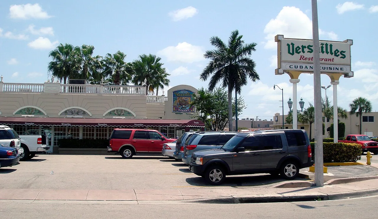 Versailles Restaurant's pylon sign reading Versailles Restaurant Cuban Cuisine in front of its cream, balustraded building with red awnings and palm trees on Calle Ocho
