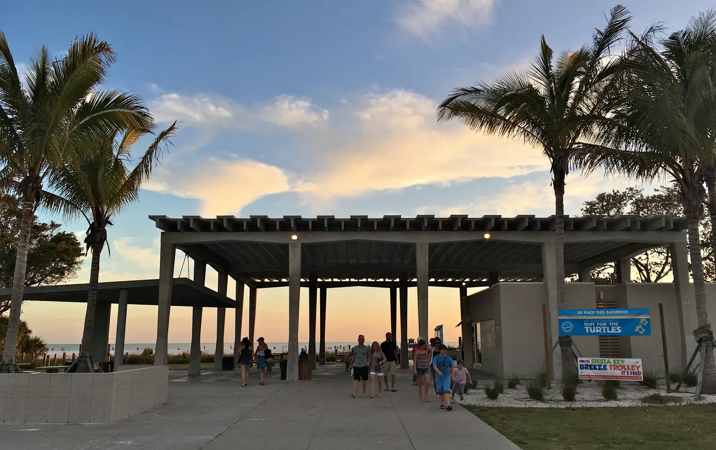 The open-air pavilion with a wood-slat roof and tall palm trees at the main Siesta Beach entrance at dusk, with visitors walking beneath it