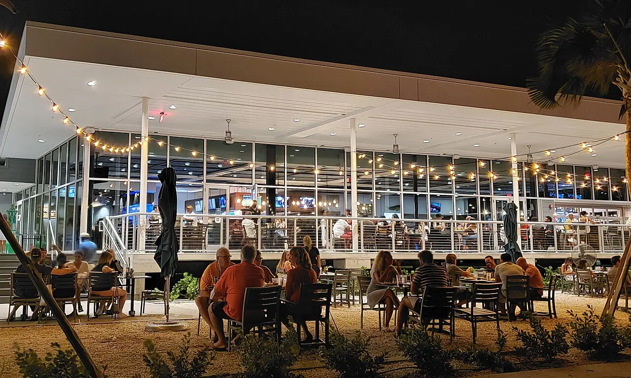 Diners at outdoor tables under string lights on the covered lower-level patio of Doc Ford's Rum Bar and Grille on the St. Petersburg Pier at night, with palm trees at the edge of the deck