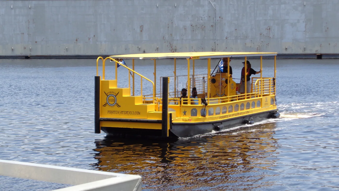 A bright yellow, open-air Pirate Water Taxi boat, its hull lettered PIRATEWATERTAXI.COM, cruising a Tampa waterway with a few passengers aboard