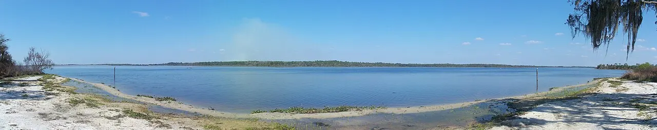 A wide panoramic view of Lake Manatee State Park's lake, calm blue water bordered by a sandy shore and a tree line under a clear sky