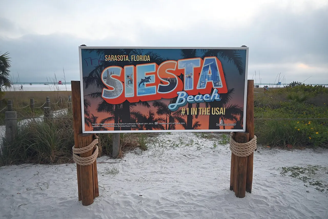 A large blue Siesta Beach entrance sign reading "Siesta Beach #1 in the USA" on a wooden post at the sandy beach entrance in Sarasota, Florida