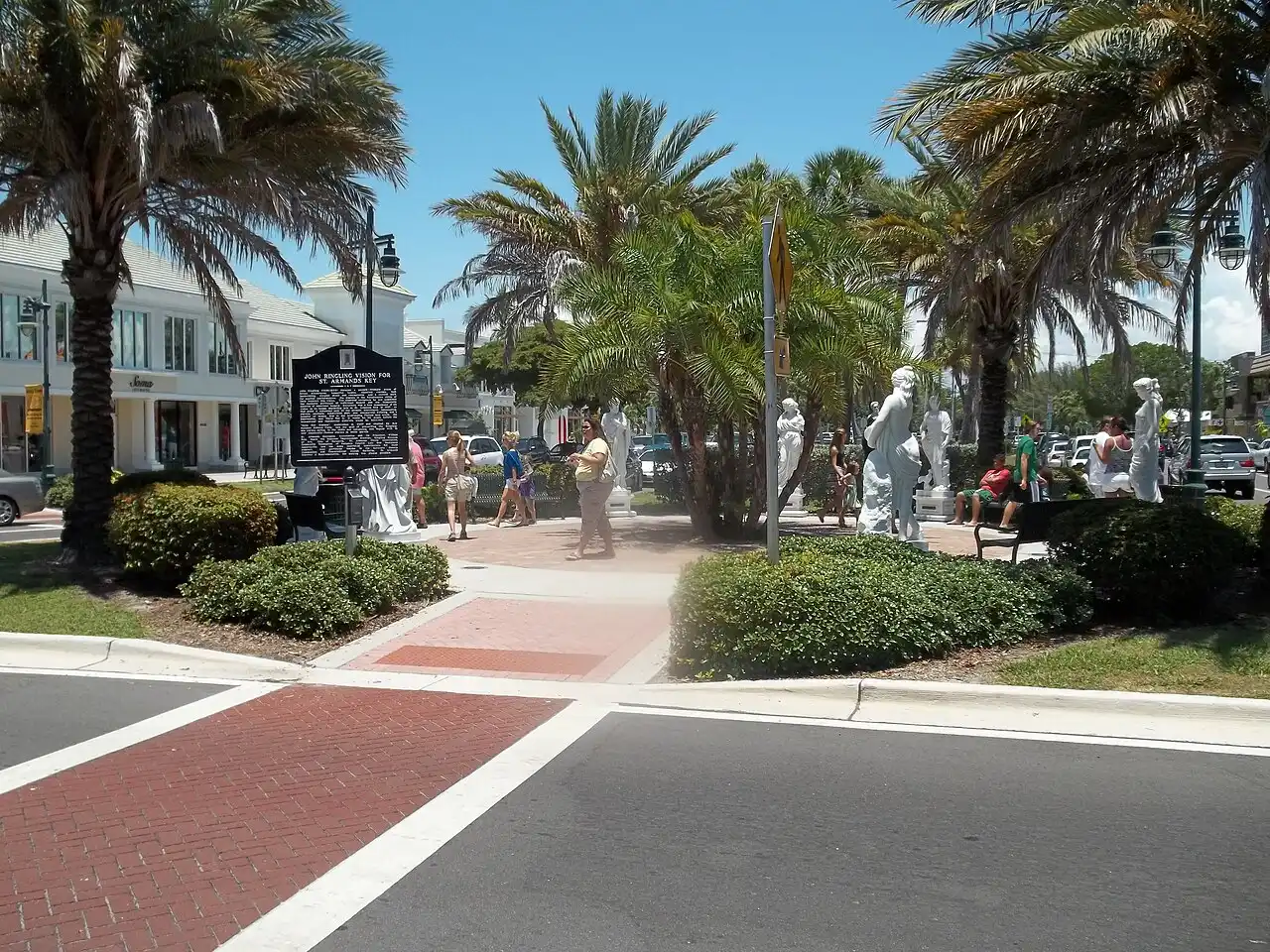 St. Armands Circle in Sarasota, with a historical marker titled "John Ringling Vision for St. Armands Key", classical statues and palm trees around the roundabout, and a few pedestrians
