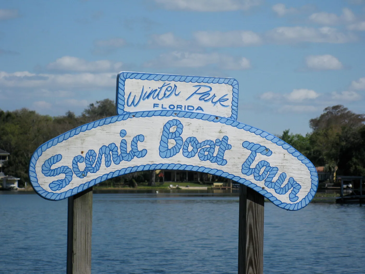 A hand-painted wooden sign reading 'Winter Park Florida Scenic Boat Tour' mounted on dock posts, with a lake behind it