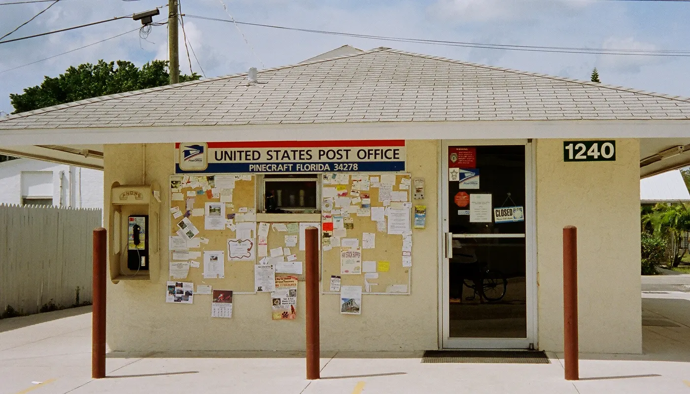 The small beige United States Post Office building in Pinecraft, with its address '1240' and a community bulletin board by the door