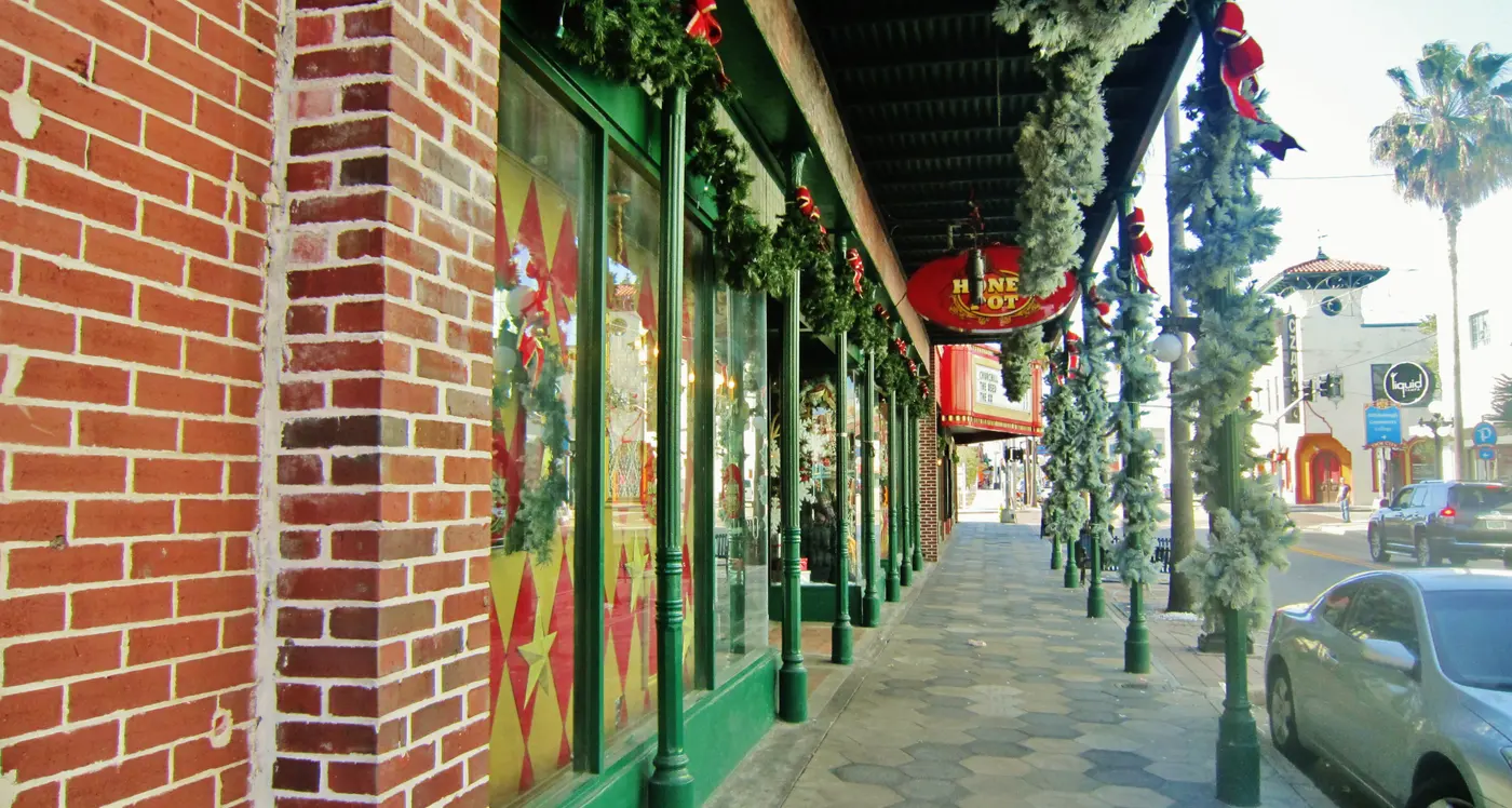 A covered sidewalk lined with shopfronts and string lights along Ybor City's Seventh Avenue, with palm trees and cars along the curb
