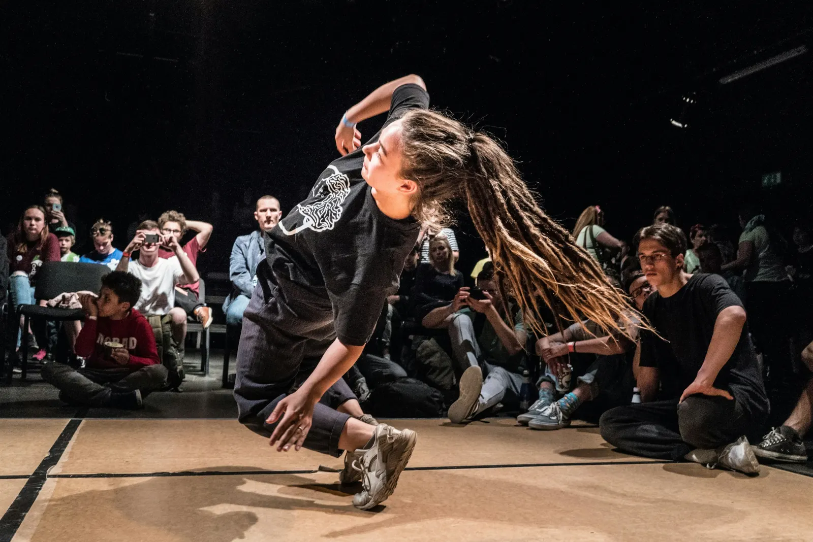 A street dancer drops low mid battle while a seated crowd watches and films