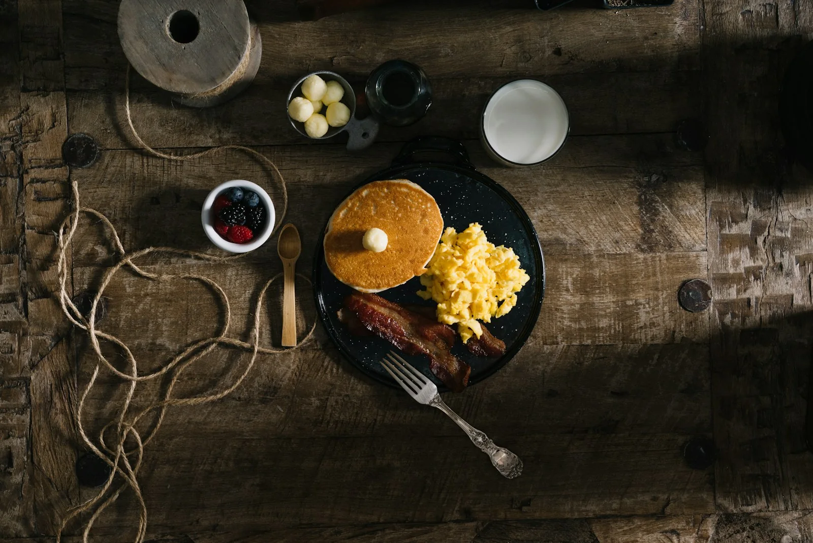 Pancakes, scrambled eggs, bacon and berries arranged on a rustic table